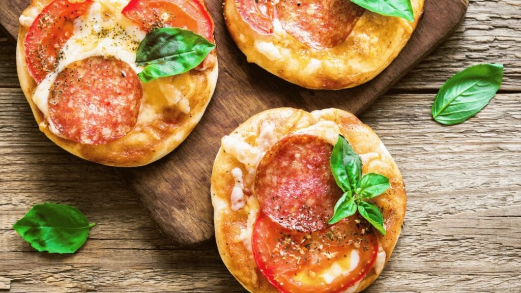 Mini pizzas individuais com queijo derretido, tomate, calabresa e folhas de manjericão.